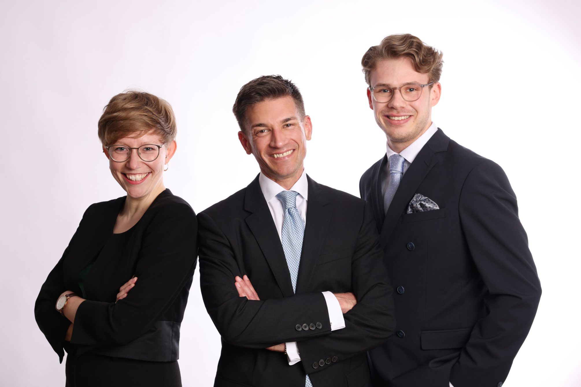 Alle Business 1 Das Bild zeigt drei Personen in formeller Geschäftskleidung, die nebeneinander stehen und freundlich in die Kamera lächeln. Links steht Marie Geffroy mit einer schwarzen Blazerjacke. In der Mitte ist Dr. André Latz, der ein schwarzes Sakko und eine hellblaue Krawatte trägt. Rechts steht Moritz Richarz, der einen dunkelblauen Anzug und eine passende Krawatte trägt. Ihre entspannten und selbstbewussten Posen, kombiniert mit ihrem warmen Lächeln, vermitteln Professionalität und Zugewandtheit.