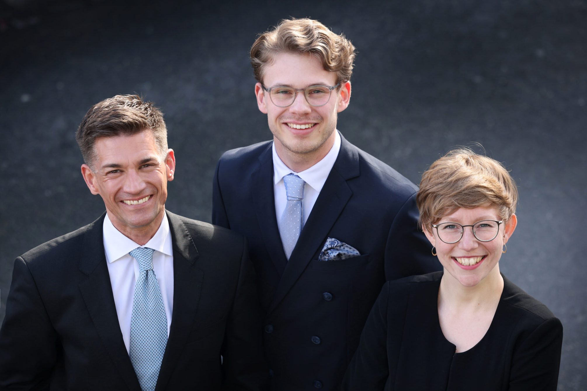 Alle Business 2 Auf dem Bild sind drei professionell gekleidete Personen abgebildet, die freundlich und zuversichtlich in die Kamera lächeln. Links steht Dr. André Latz in einem dunklen Anzug und heller Krawatte, in der Mitte Moritz Richarz in einem dunkelblauen, zweireihigen Anzug, und rechts Marie Geffroy, die einen eleganten schwarzen Anzug trägt. Ihre freundlichen Gesichtsausdrücke und die formelle Kleidung vermitteln Kompetenz und Professionalität.