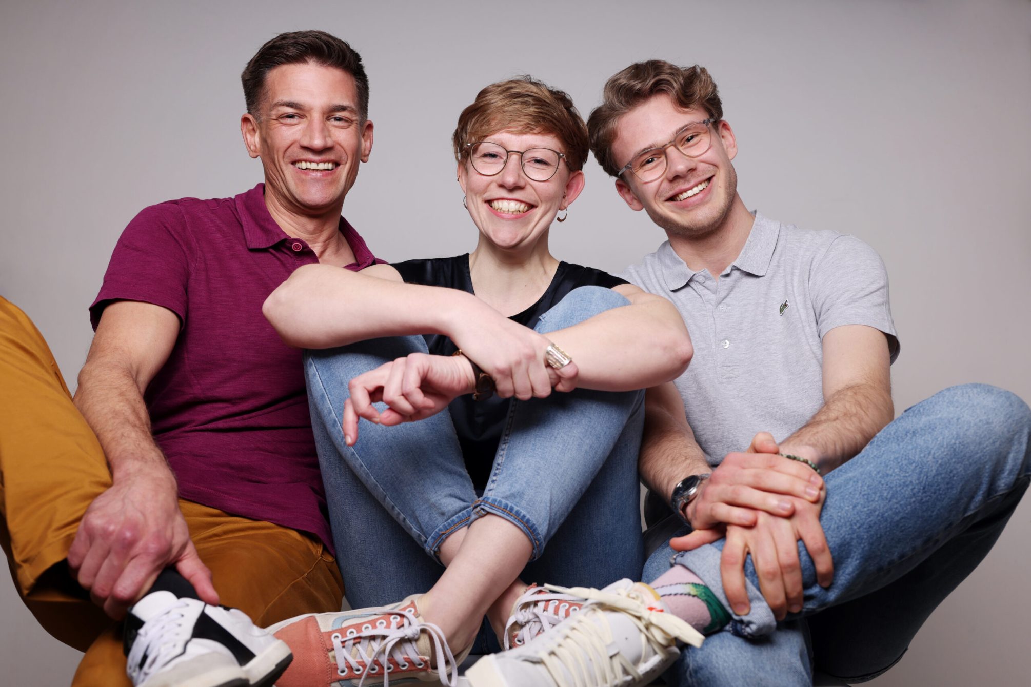 Alle casual Gruppenfoto: Das Bild zeigt Dr. André Latz, Marie Geffroy und Moritz Richarz, die ungezwungen und entspannt zusammen sitzen. Sie wirken fröhlich und energiegeladen und tragen legere Kleidung und verschiedene Accessoires, die ihre Persönlichkeit unterstreichen. Ihre ausgestreckten Arme und verschlungenen Hände symbolisieren Einheit und Teamgeist. Ihr freundliches Lächeln und die direkte Kameraposition zeigen Offenheit und Zugänglichkeit. Sie präsentieren sich als kompetentes und dynamisches Team, das Professionalität mit einer entspannten Atmosphäre verbindet.