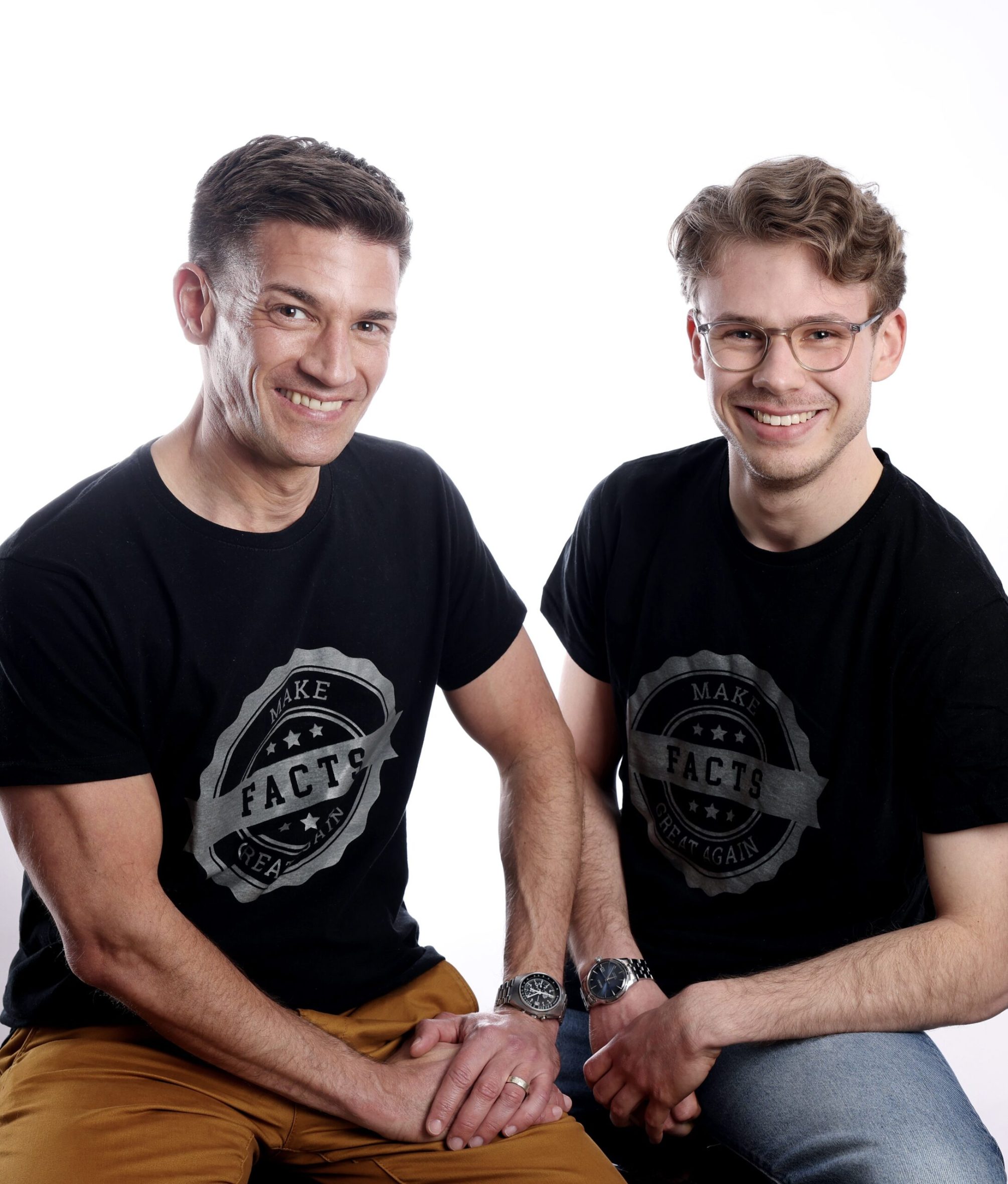 Andre Moritz Facts Foto von André Latz und Moritz Richarz
André Latz (links) und Moritz Richarz (rechts) sitzen nebeneinander und lächeln in die Kamera. Beide tragen schwarze T-Shirts mit der Aufschrift „Make Facts Great Again“ in grauer Schrift auf einem runden Emblem. André Latz trägt braune Hosen, Moritz Richarz eine Jeans. Beide haben Uhren am Handgelenk. Der Hintergrund ist einfarbig weiß.