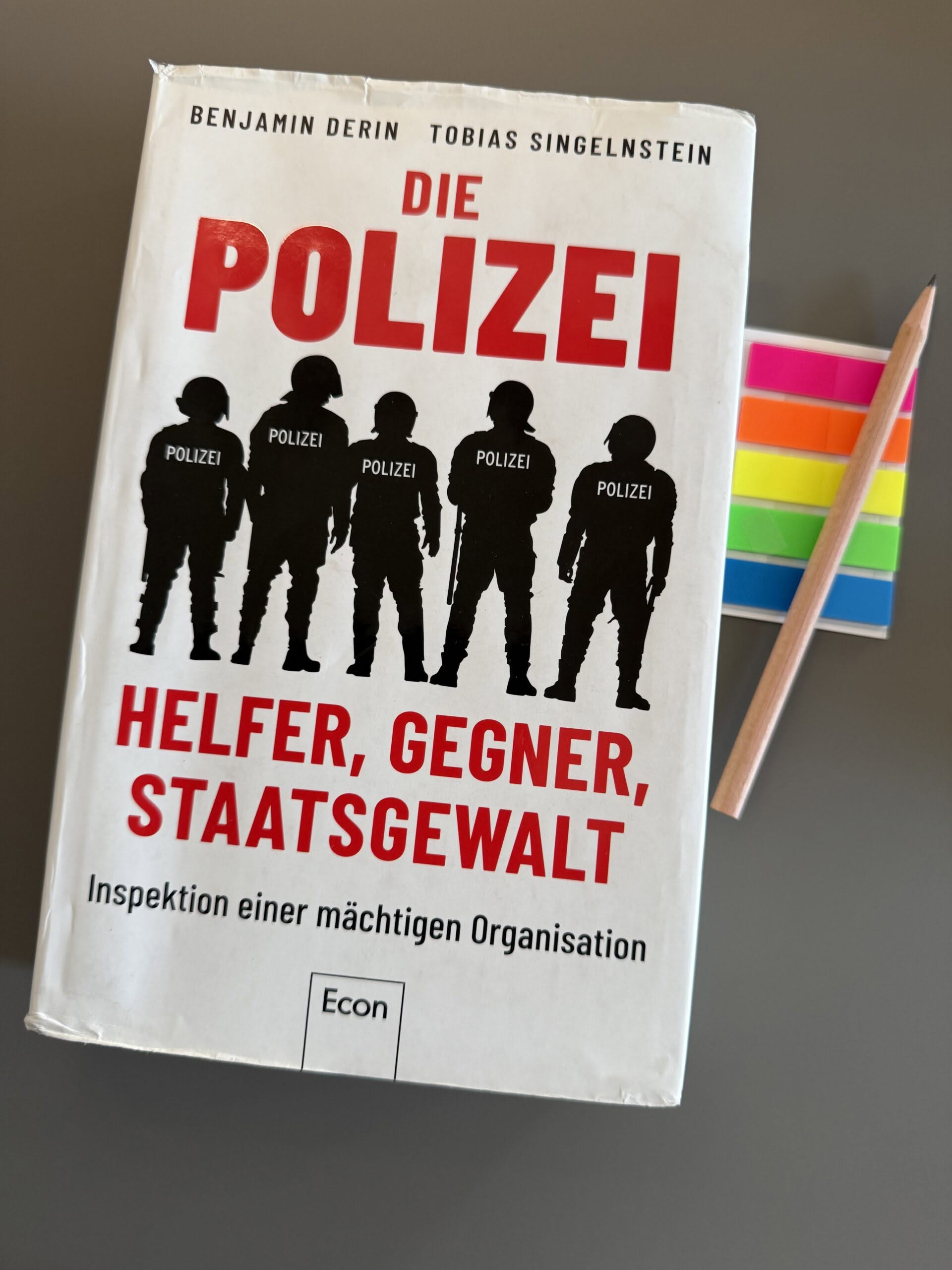 Cover Derin-Singelnstein_Polizei_2 Das Buch "Derin, Benjamin / Singelnstein, Tobias (2022): Die Polizei. Helfer, Gegner, Staatsgewalt" auf grauem Untergrund. Rechts neben dem Buch liegen ein Bleistift und ein Briefchen bunter Markierungsklebestreifen.