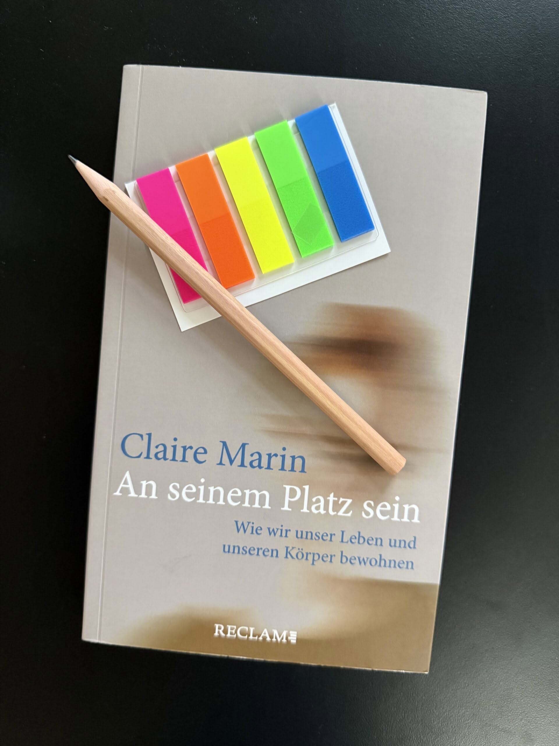 Cover Marin_An_Platz_sein_2 Das Buch "Marin, Claire (2023): An seinem Platz sein. Wie wir unser Leben und unseren Körper bewohnen" auf schwarzem Untergrund. Auf dem Buch liegen ein Bleistift und ein Briefchen bunter Markierungsklebestreifen.
