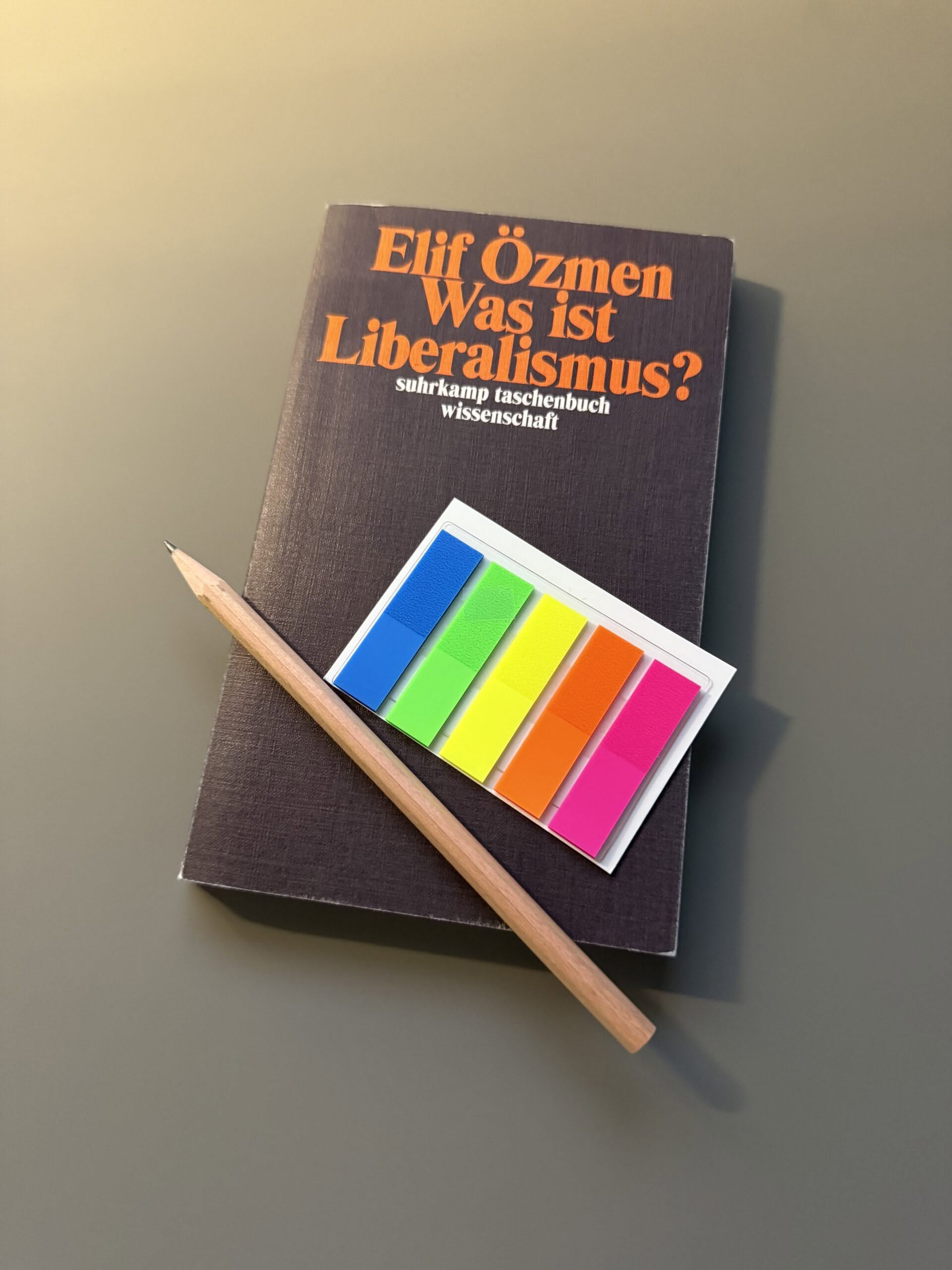Cover Oezmen_was-ist-liberalismus_2 Das Buch "Özmen, Elif (2023): Was ist Liberalismus?" auf grauem Untergrund. Auf dem Buch liegen ein Bleistift und ein Briefchen bunter Markierungsklebestreifen.