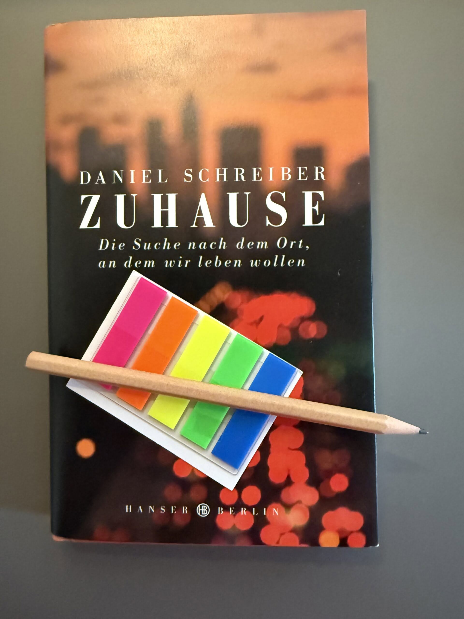 Cover Schreiber_Zuhasue_2 Das Buch "Schreiber, Daniel (2022): Zuhause. Die Such nach dem Ort, an dem wir leben wollen. 4. Aufl. Hanser (Berlin)." auf grauem Untergrund. Auf dem Buch liegen ein Bleistift und ein Briefchen bunter Markierungsklebestreifen.