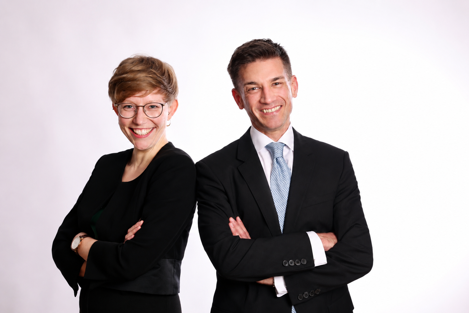 Zwei lächelnde Personen in Businesskleidung stehen mit verschränkten Armen vor hellem Studiohintergrund.
