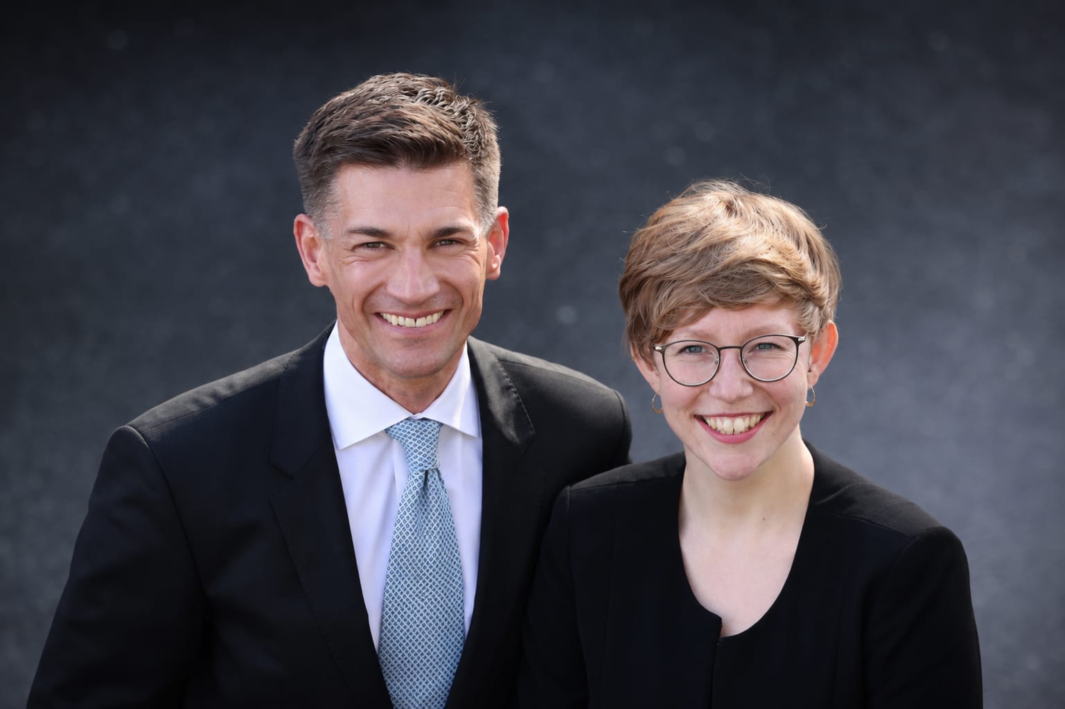 Business-Porträt vor dunklem Hintergrund Zwei lächelnde Personen in dunkler Businesskleidung stehen nah beieinander vor dunkel verschwommenem Hintergrund.